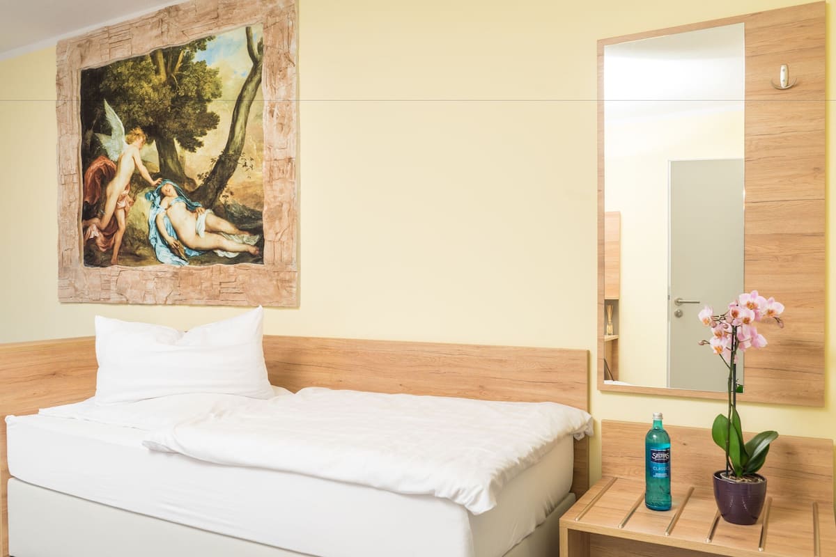 A hotel room with a bed, a wooden headboard, a painting, a mirror, a nightstand, a bottle, and a potted orchid.