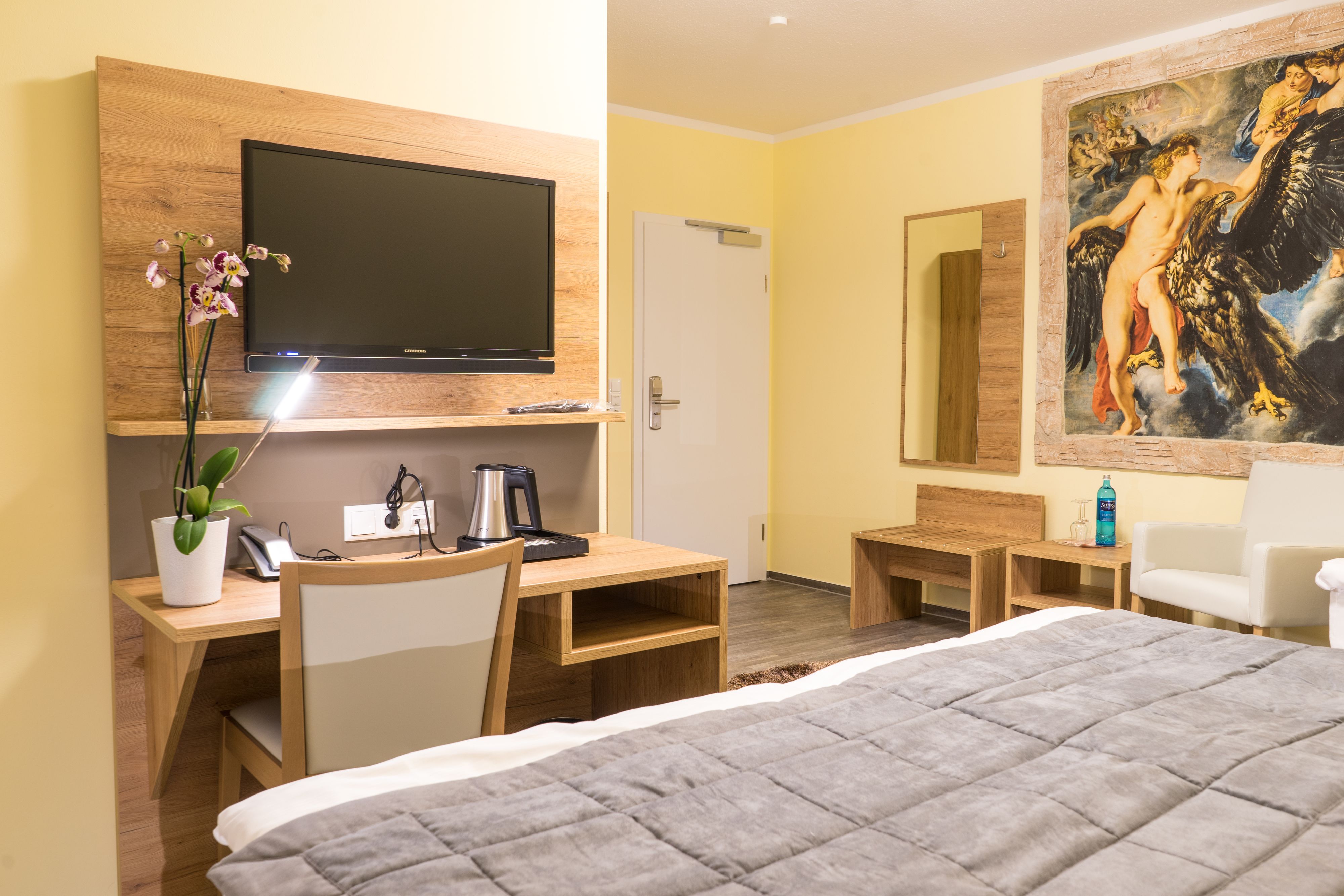 A hotel room with a bed, a desk with a chair, a television, a painting, and a door.