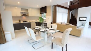 A modern kitchen with white cabinets, a dining table with chairs, and a living area with a sofa and a TV. - AnB Pool Villa 3BR Glass House in Pattaya (Sattahip)