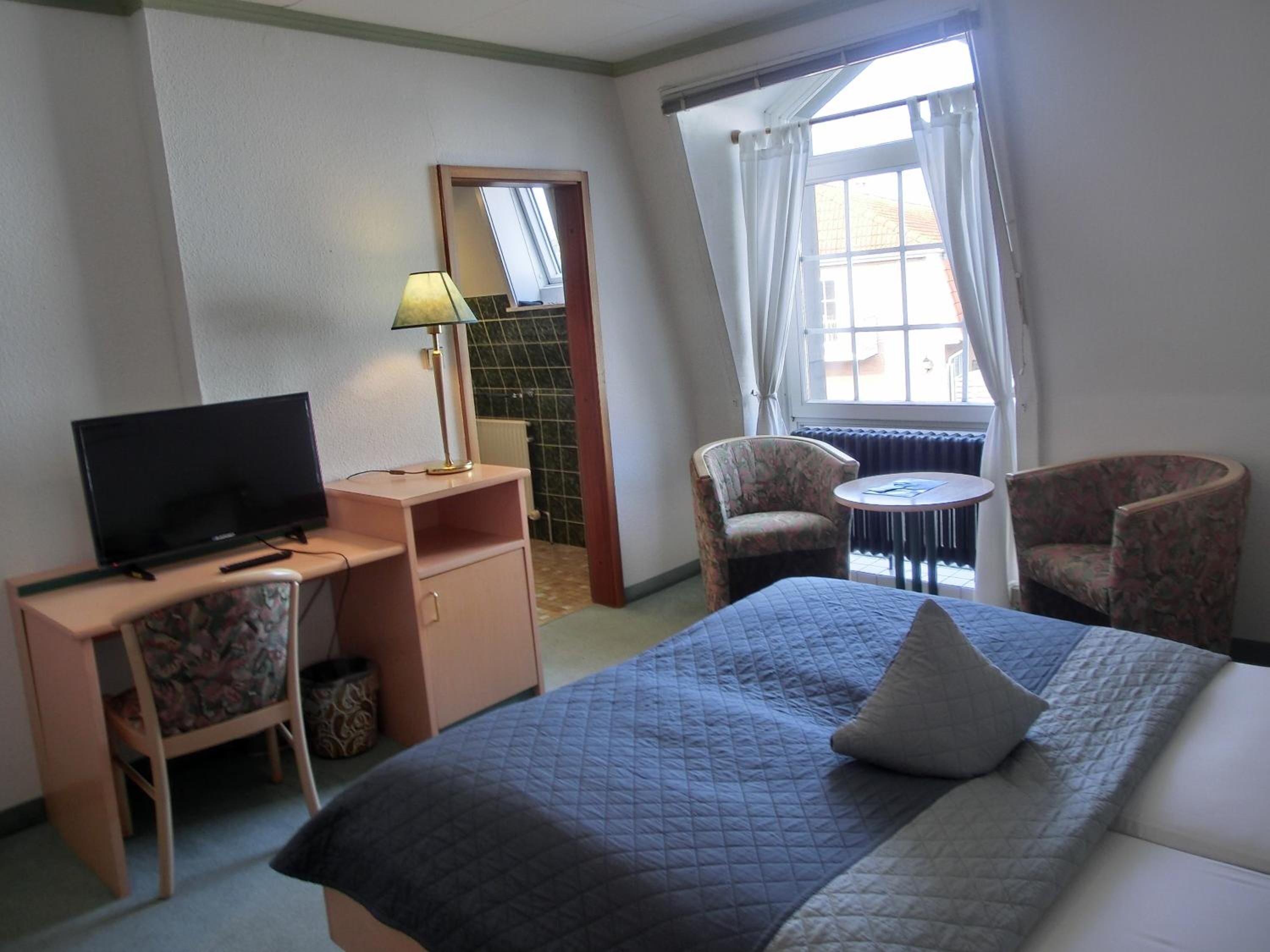 A hotel room with a bed, a desk with a television, a chair, and a small table.