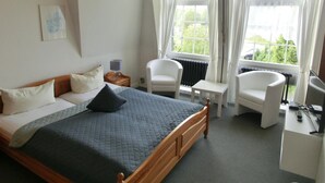 Une chambre d’hôtel avec un lit, deux chaises blanches, une petite table et un bureau équipé d’un ordinateur et d’un téléphone.