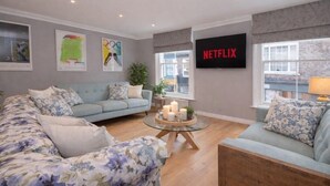 Un salón con un sofá azul claro, un sofá estampado con motivos florales, una mesa de centro de cristal con velas y un televisor de pared mostrando 'NETFLIX'.