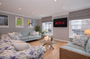 Ein Wohnzimmer mit einem hellblauen Sofa, einem Sofa mit Blumenmuster, einem Glastisch mit Kerzen und einem an der Wand montierten Fernseher, der 'NETFLIX' anzeigt.