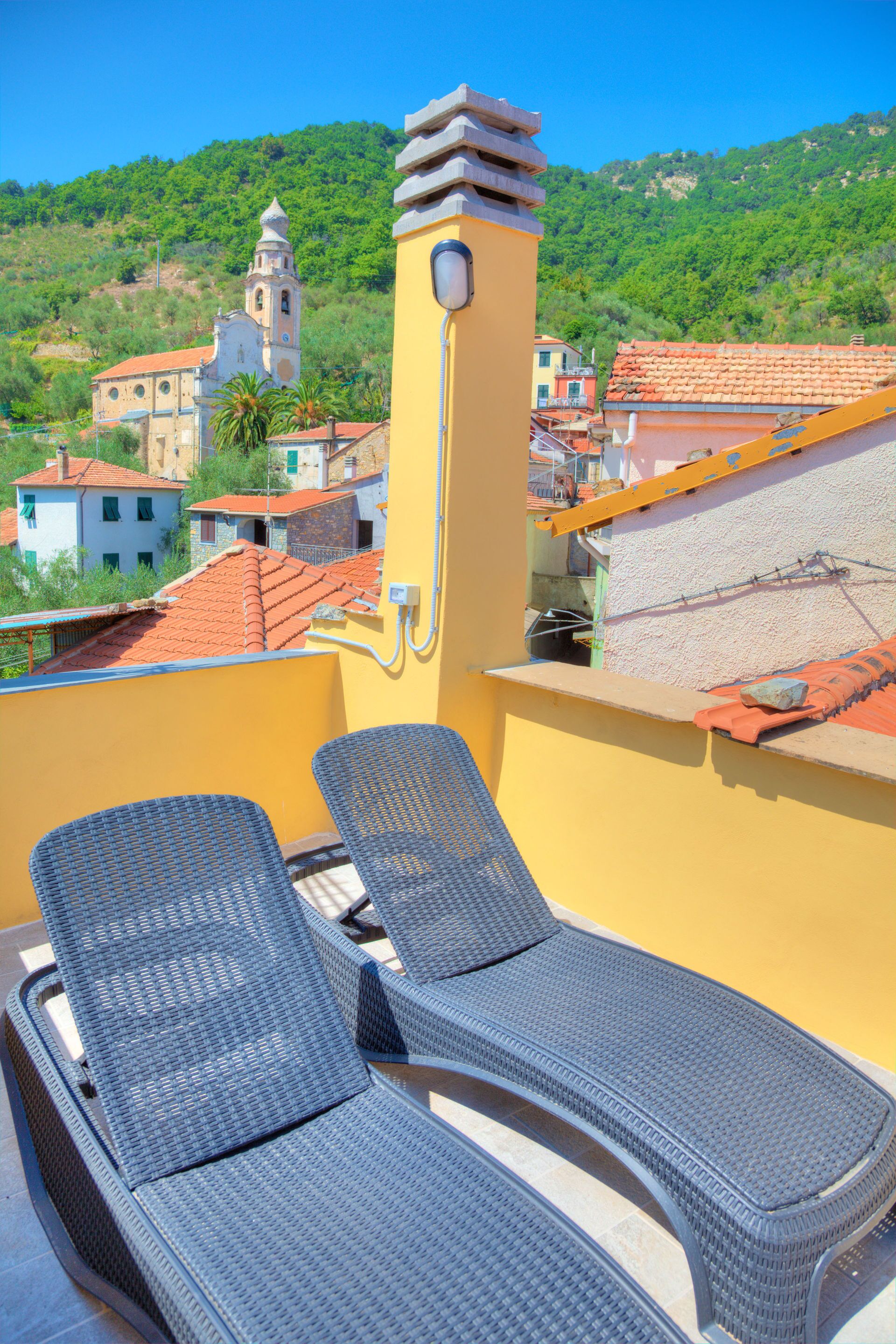 A rooftop terrace with two lounge chairs, a yellow wall, and a view of a town with buildings and a hill in the background.