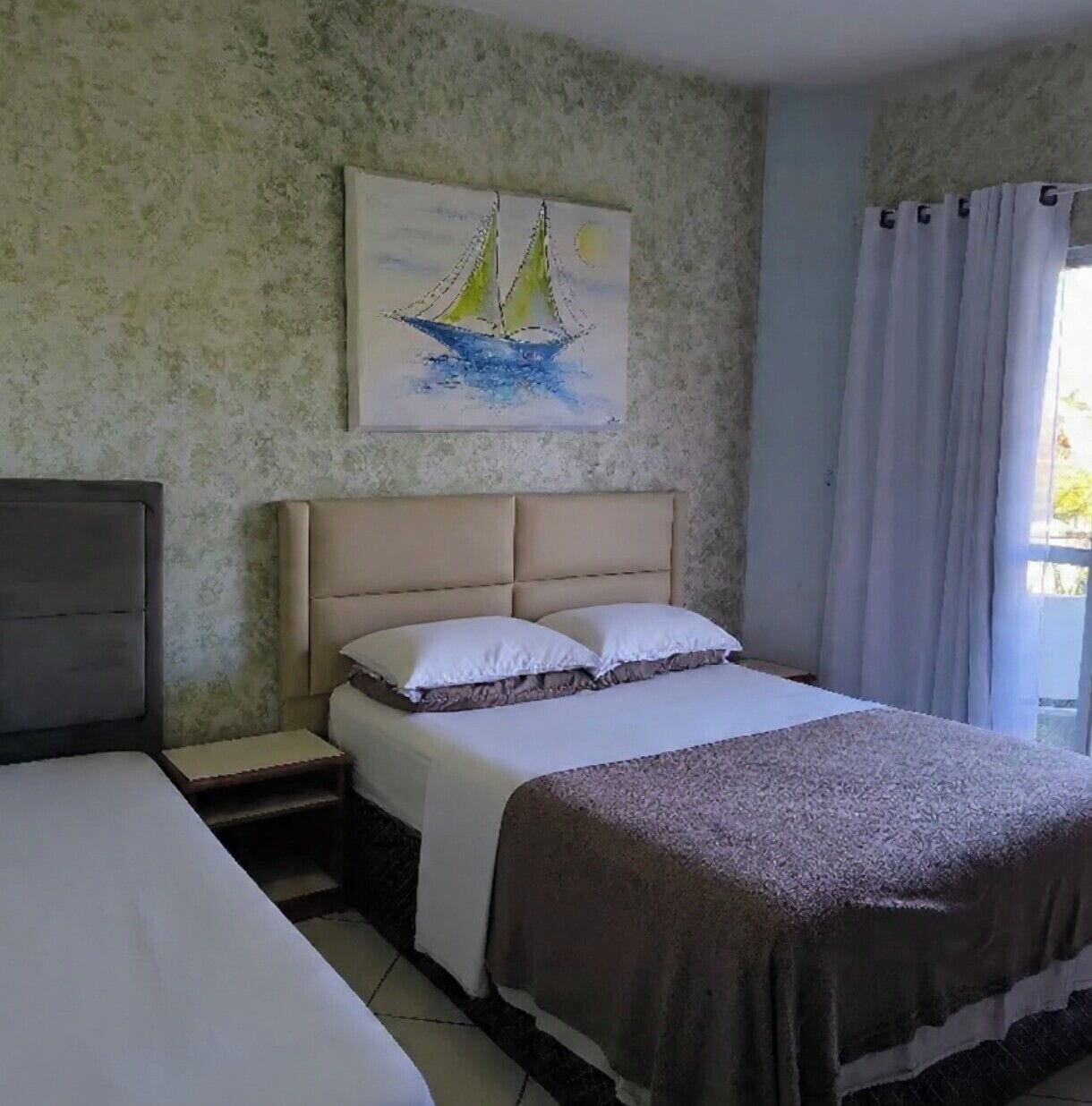 Un dormitorio con cama, mesita de noche, cabecera, un cuadro de veleros y una ventana con cortinas.