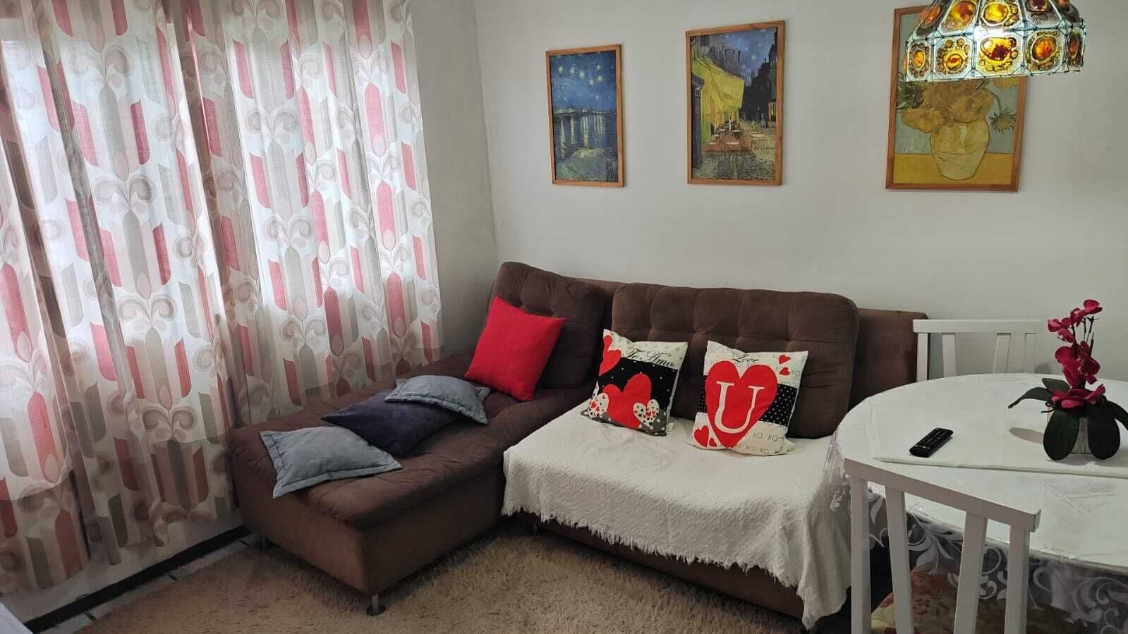 Un salón con un sofá marrón, una mesa blanca y un cojín rojo. Hay tres cuadros en la pared.