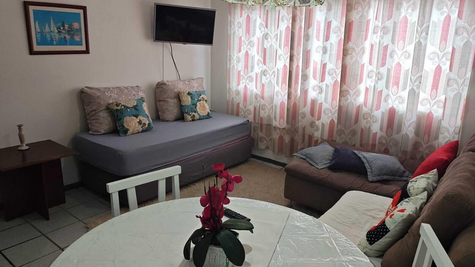 A living room with a sofa, a coffee table with a vase, a TV mounted on the wall, and a patterned curtain.