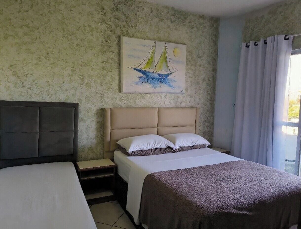 Un dormitorio con cama, mesita de noche, cabecera, un cuadro de un velero y una ventana con cortinas.