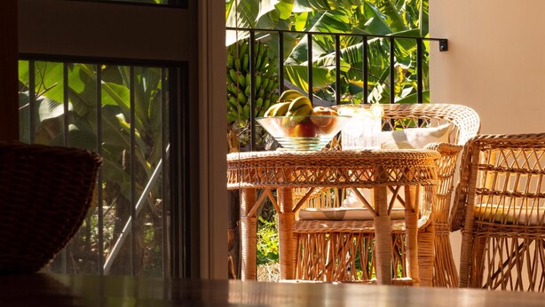 A wicker table with a glass bowl of fruit, a wicker chair, and a view of lush green foliage through a window with a metal railing.