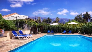 A swimming pool with blue water, surrounded by lounge chairs and umbrellas, with mountains in the background.