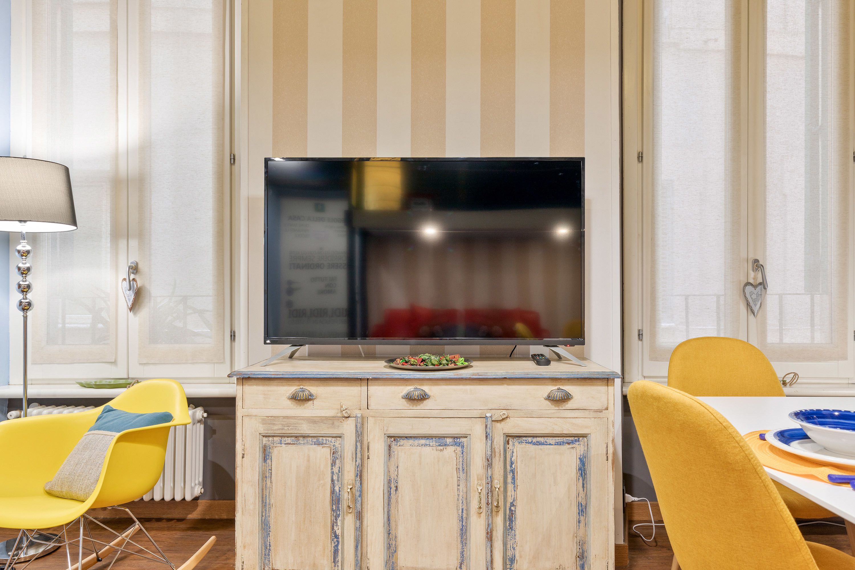 Una habitación con una televisión sobre un mueble, sillas amarillas y una mesa con un bol.
