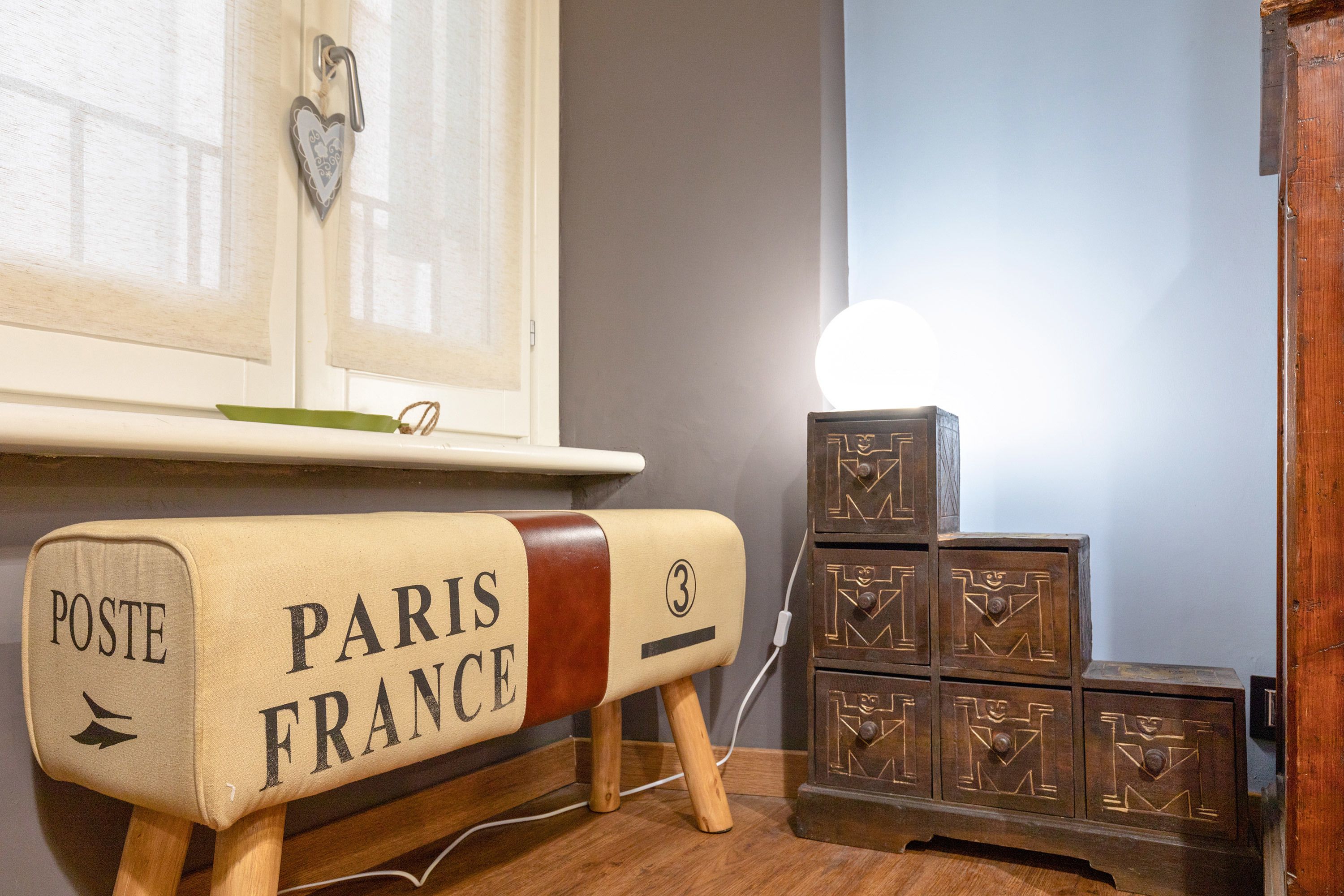 A vintage-style mailbox with "POSTE PARIS FRANCE" on it, wooden cabinets with geometric patterns, and a window with sheer curtains.