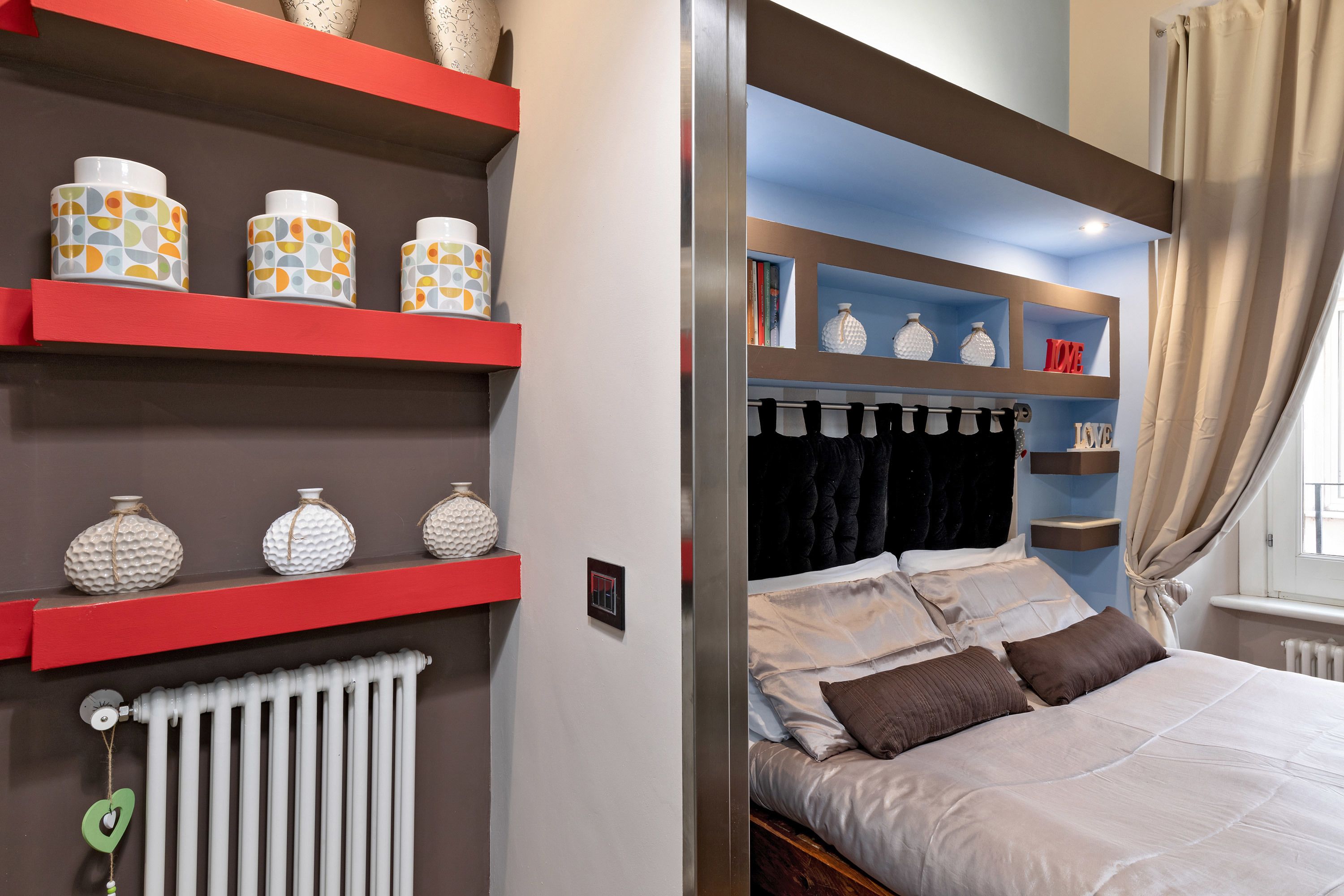 A bedroom with a bed, red shelving with decorative items, and a window with curtains.