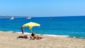 Uma praia com um guarda-sol amarelo, pessoas tomando sol e barcos na água.