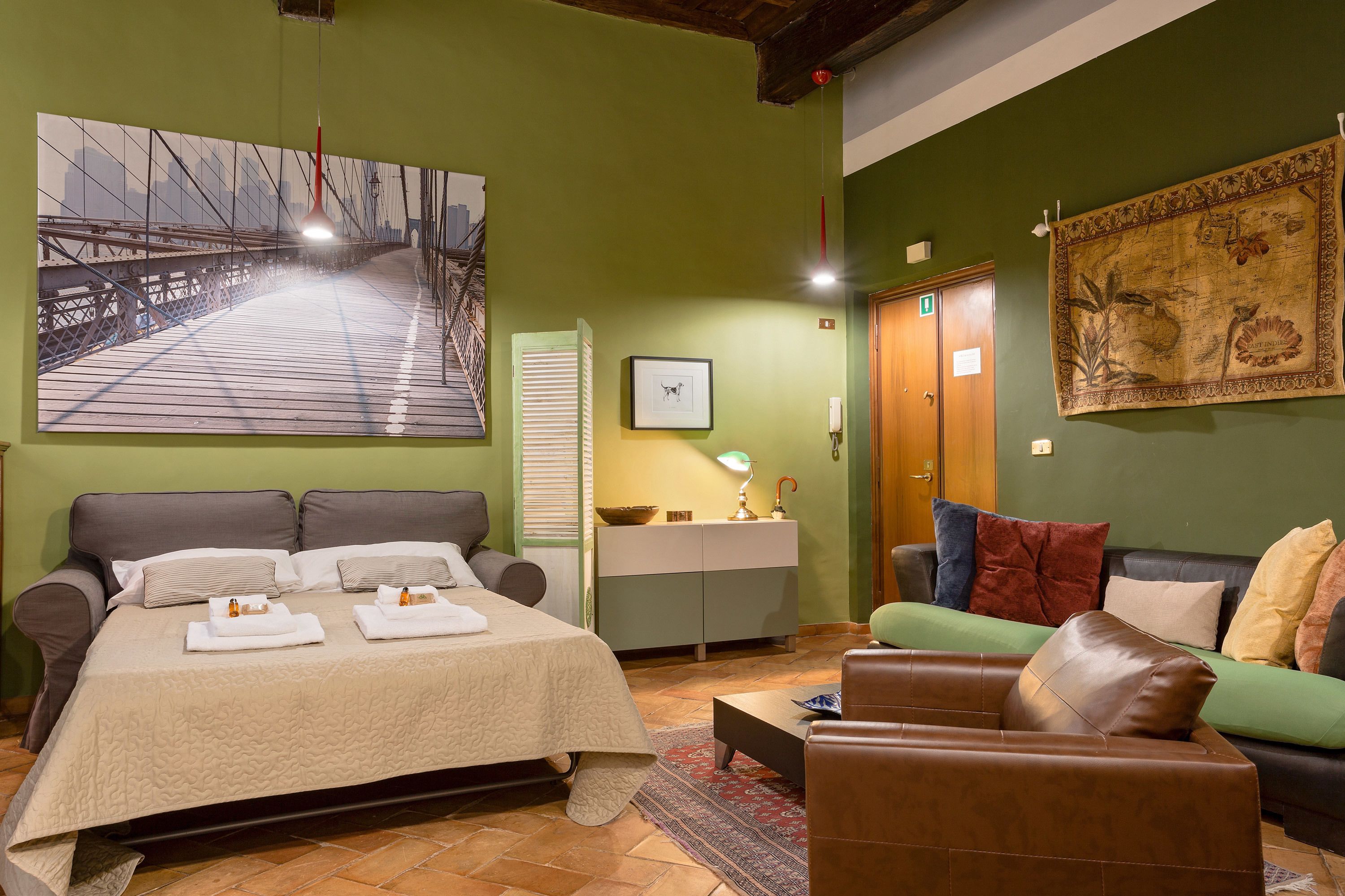 A bedroom with a large bed, a green sofa, a brown leather chair, a small table, and a painting of a bridge.