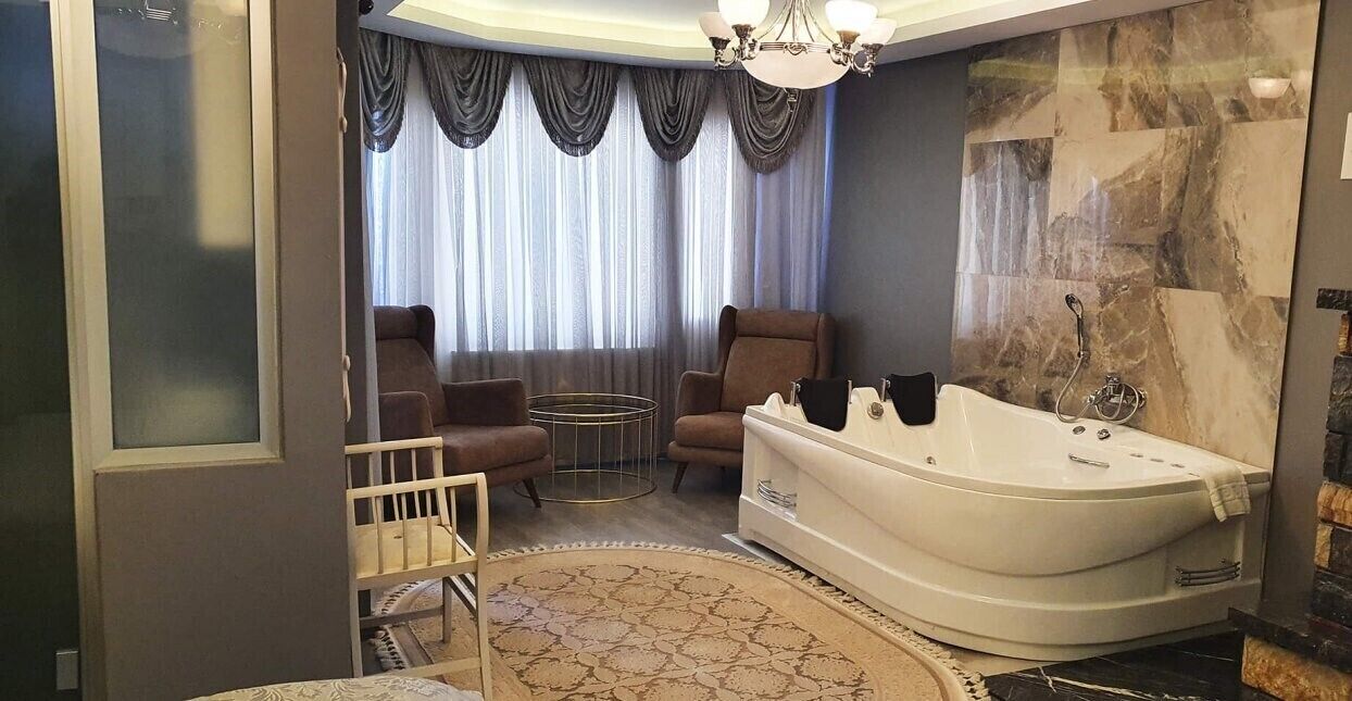 A modern bathroom with a freestanding bathtub, two armchairs, a round table, and a large window with curtains.