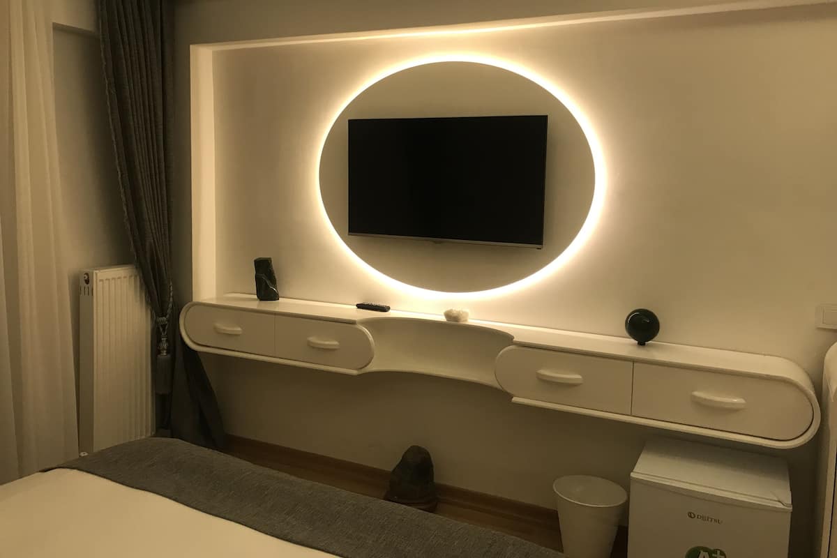 Habitación de hotel moderna con una cama grande, una mesita de noche, un televisor de pantalla plana montado en la pared con una luz circular, un cubo de basura pequeño y un radiador.