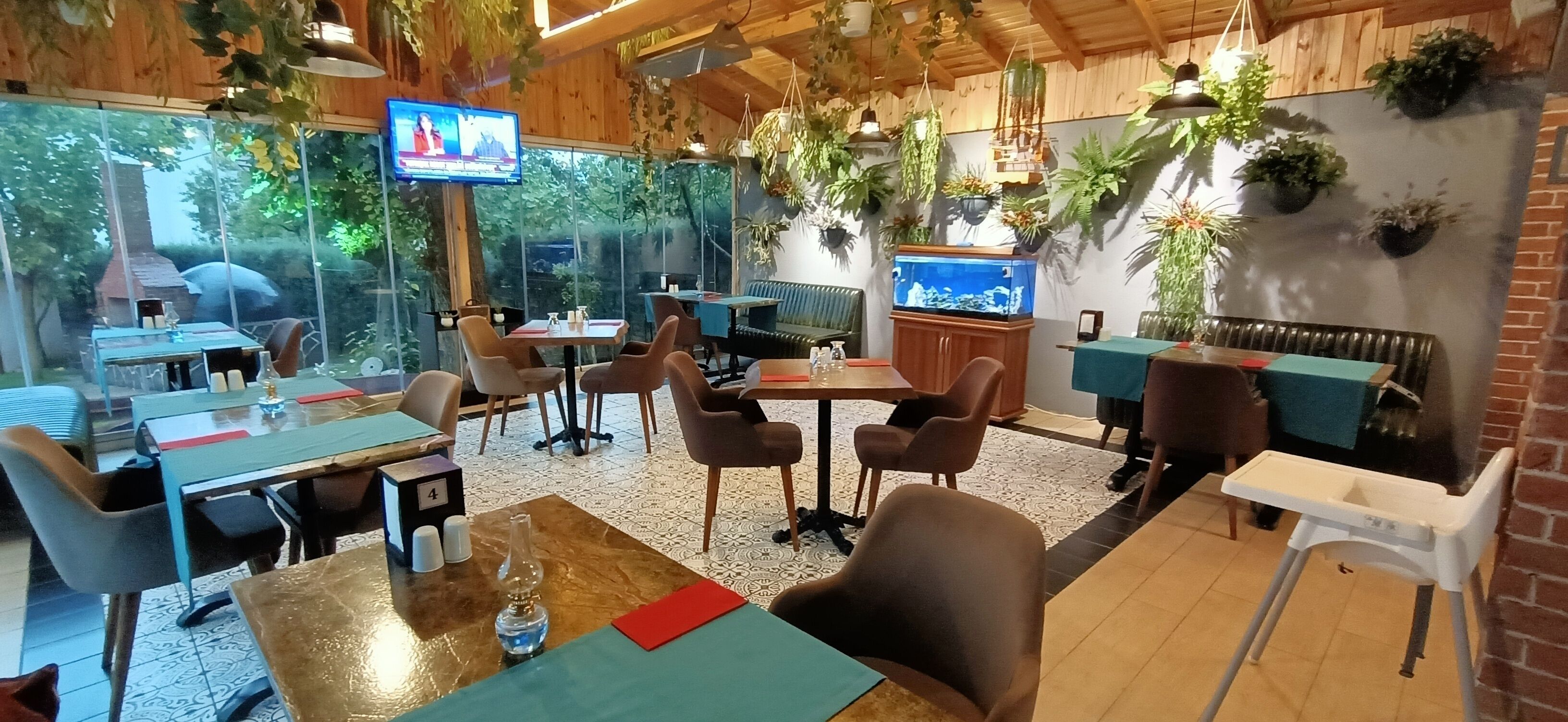 En hyggelig restaurant med trævægge, hængende planter og et tv-skærm. Der er borde og stole stillet frem til spisning.