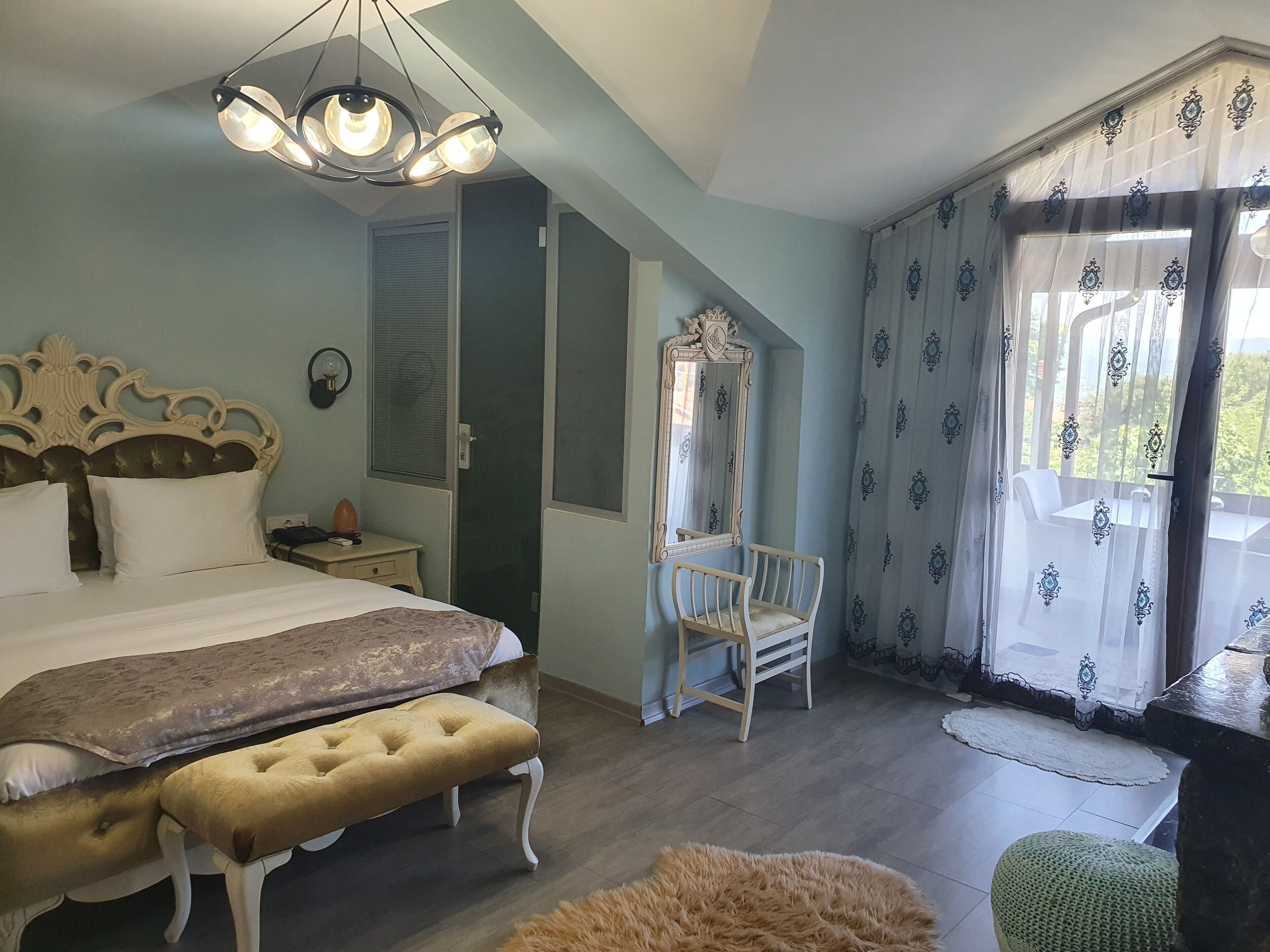 Un dormitorio con cama, un banco, una silla, un espejo y una ventana con cortinas.