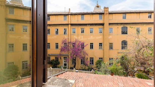 A courtyard with a yellow building, a wooden staircase, and a tree with purple flowers. - Rome as you feel - Cappellari 35 (Rome)