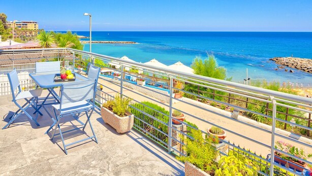 Un balcon avec une table et des chaises, donnant sur une mer bleue et limpide et une côte rocheuse.