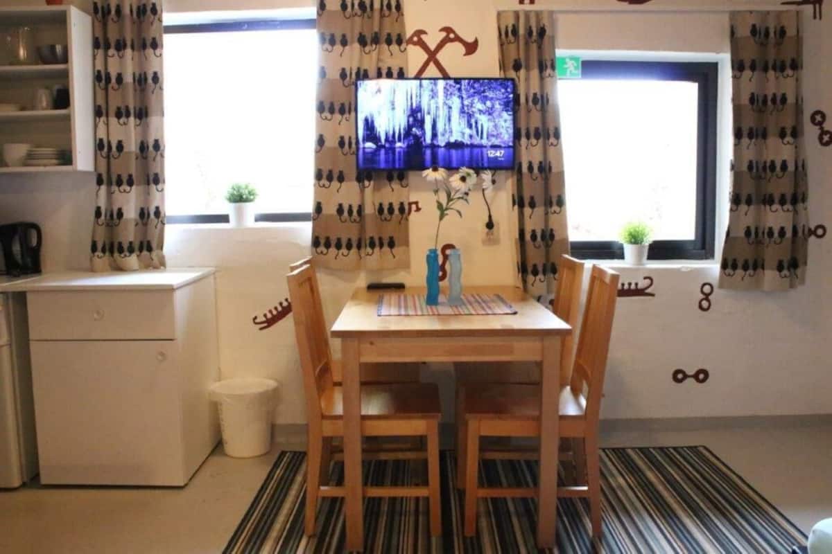 Une petite pièce agréable, avec une table et des chaises en bois, un téléviseur fixé au mur, une petite plante en pot et une fenêtre avec des rideaux.