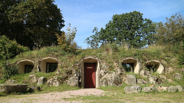 A stone structure with a central red door and three smaller arched openings, surrounded by rocks and vegetation.