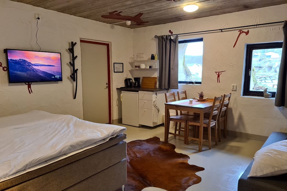 Une pièce confortable avec un lit, une table en bois et des chaises, un petit coin cuisine et un téléviseur fixé au mur.