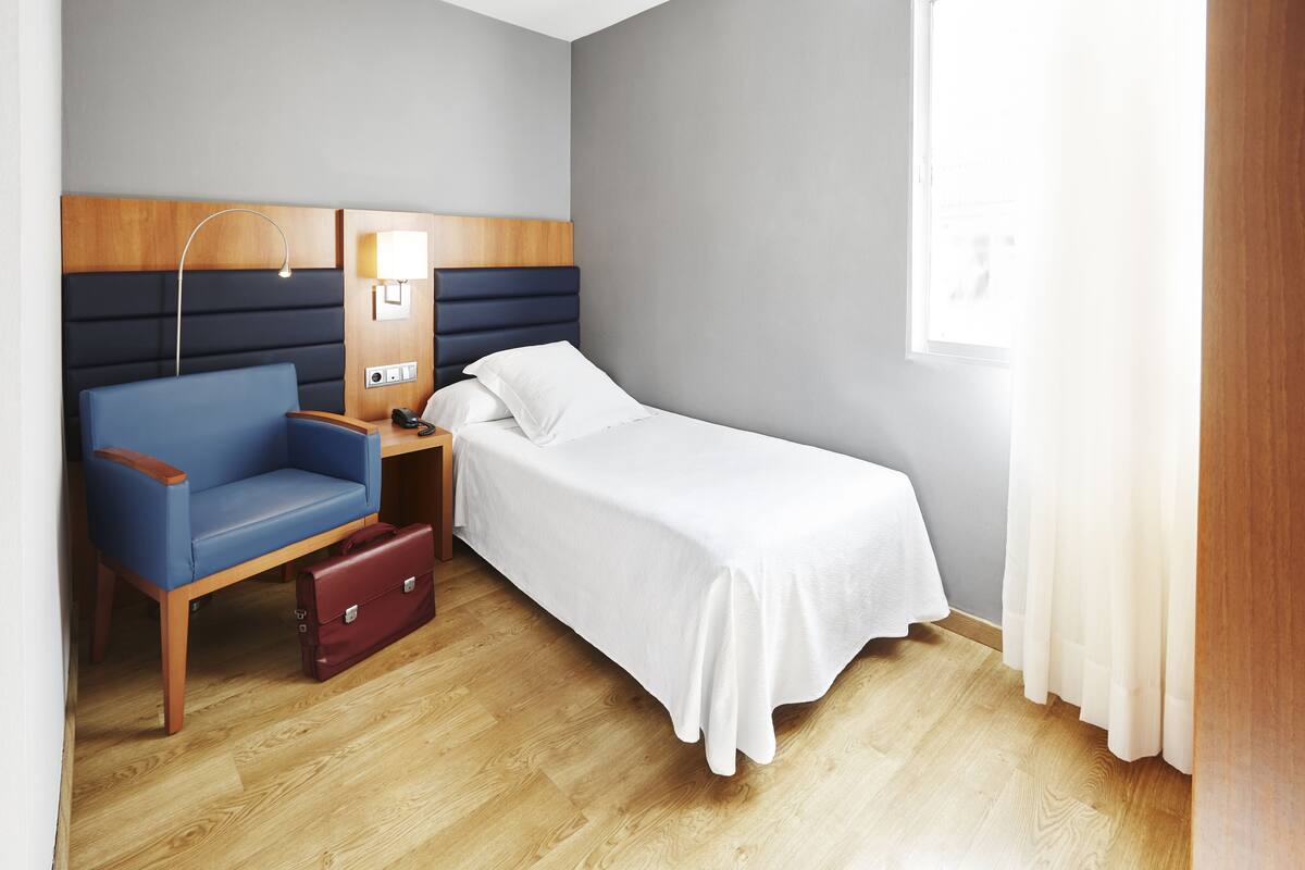 Hotellihuone, jossa on yksi sänky, puinen yöpöytä, sininen tuoli, punainen matkalaukku ja sängyn vieressä oleva lamppu.