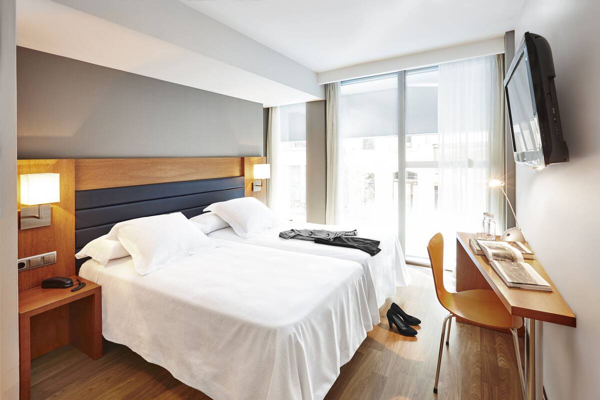 Hotellihuone, jossa on suuri sänky, työpöytä tuolilla, seinälle asennettu televisio ja ikkuna, josta on näkymä rakennuksiin.