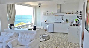 Une cuisine moderne avec des meubles blancs, un coin repas doté d’une table et de chaises blanches, et une vue sur la mer grâce à de grandes fenêtres.