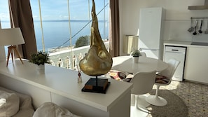 A modern kitchen with a view of the sea, a dining area with white chairs, and a unique gold sculpture on the counter.