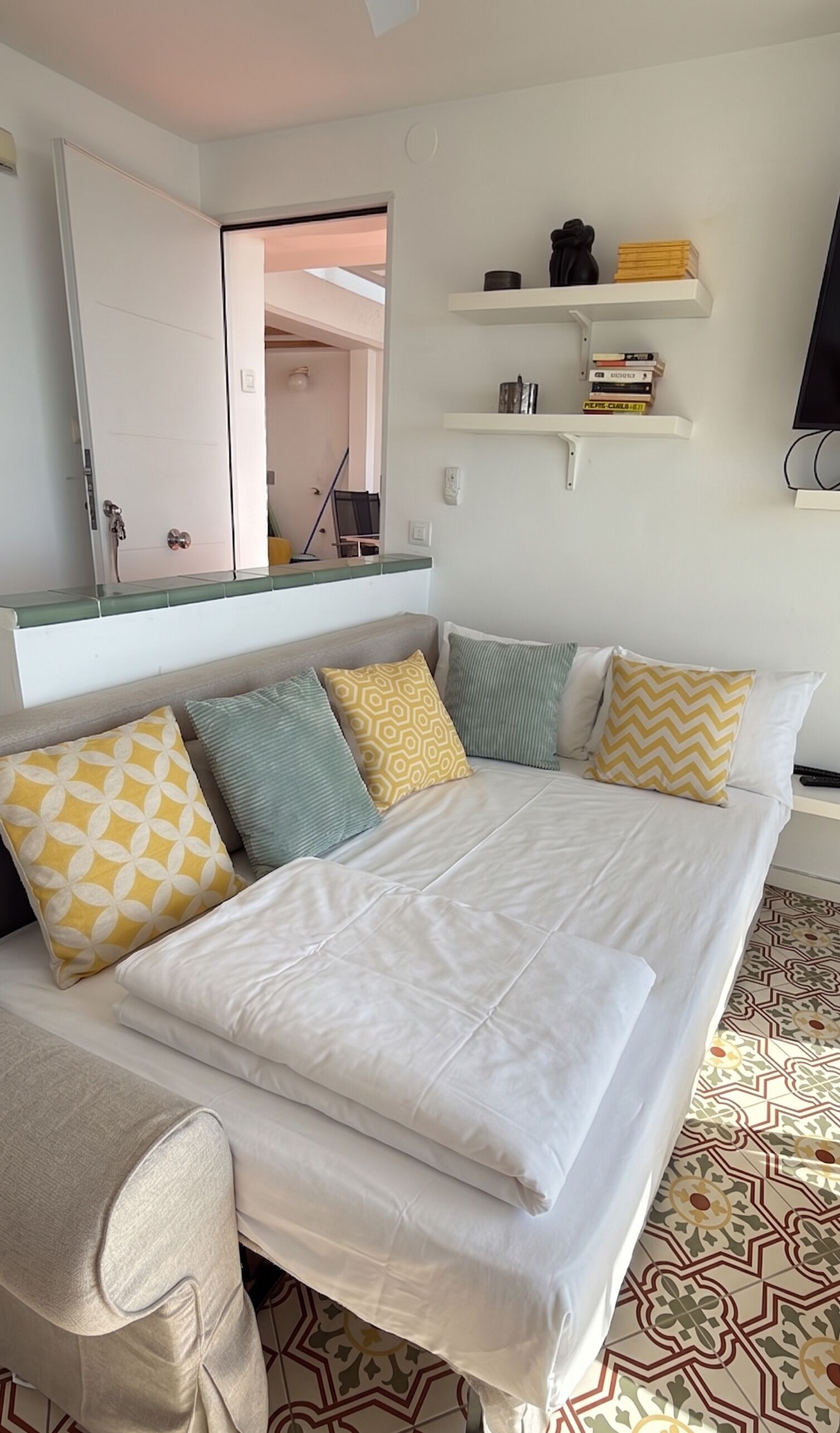 Una cama con ropa de cama blanca, una mesita de noche, un espejo y una estantería con libros y objetos decorativos.