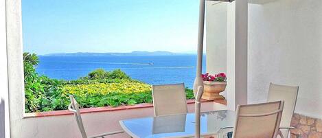 Un balcon avec une table et des chaises, un pot de fleurs et une vue sur la mer et la verdure.
