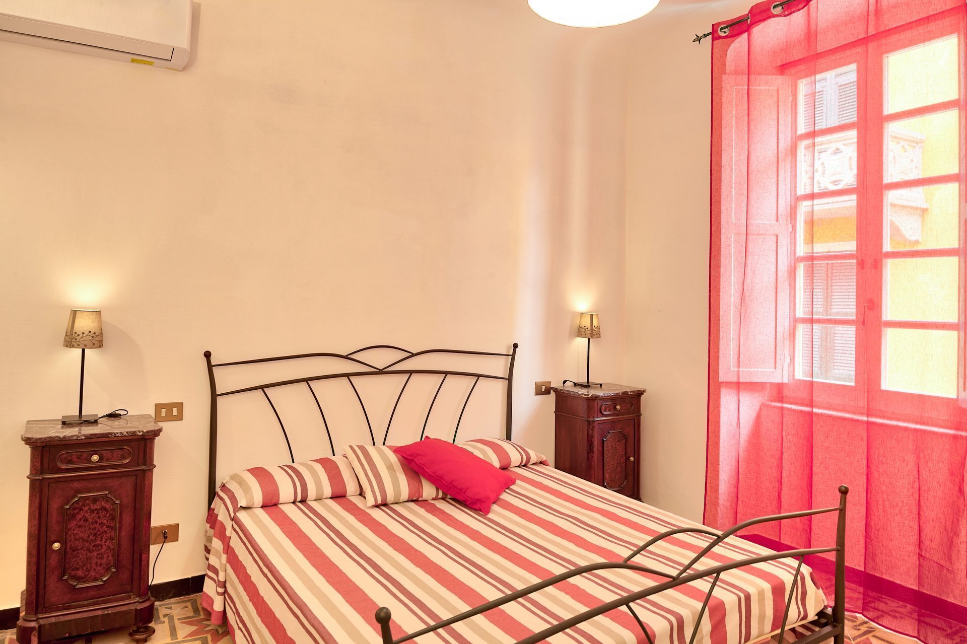 Un lit simple, recouvert d’une couvre-lit à rayures et agrémenté d’un oreiller rouge, est placé de part et d’autre de deux tables de chevet sur lesquelles reposent des lampes. La pièce est dotée d’une fenêtre ornée d’un rideau rouge.