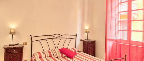 Un lit simple, recouvert d’une couvre-lit à rayures et agrémenté d’un oreiller rouge, est placé de part et d’autre de deux tables de chevet sur lesquelles reposent des lampes. La pièce est dotée d’une fenêtre ornée d’un rideau rouge.