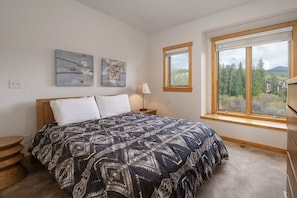 Quarto com cama grande, duas obras de arte penduradas na parede, janela com vista para árvores e montanhas, e mesa de cabeceira de madeira.