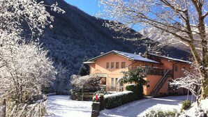 Um edifício coberto de neve com uma escadaria que leva a uma varanda, rodeado por árvores carregadas de neve e uma montanha ao fundo.