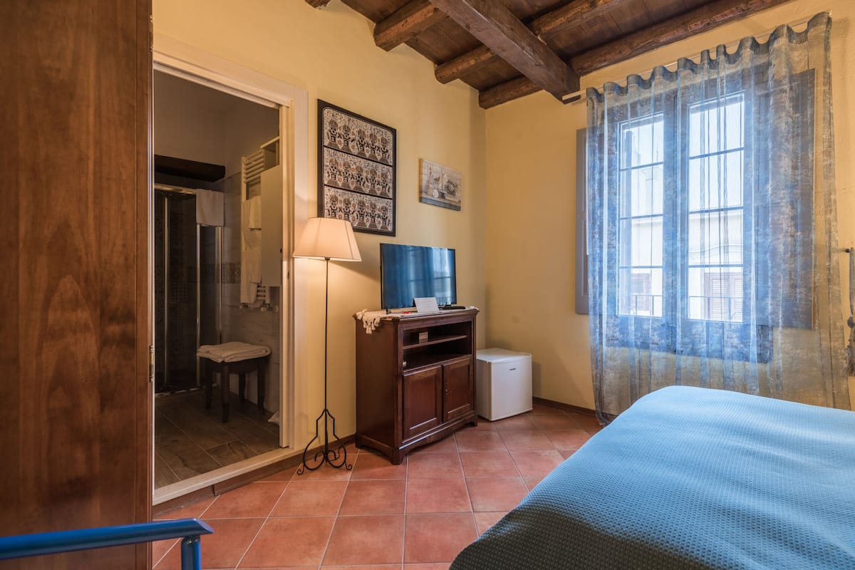 A bedroom with a bed, a small cabinet, a television, and a window with curtains.
