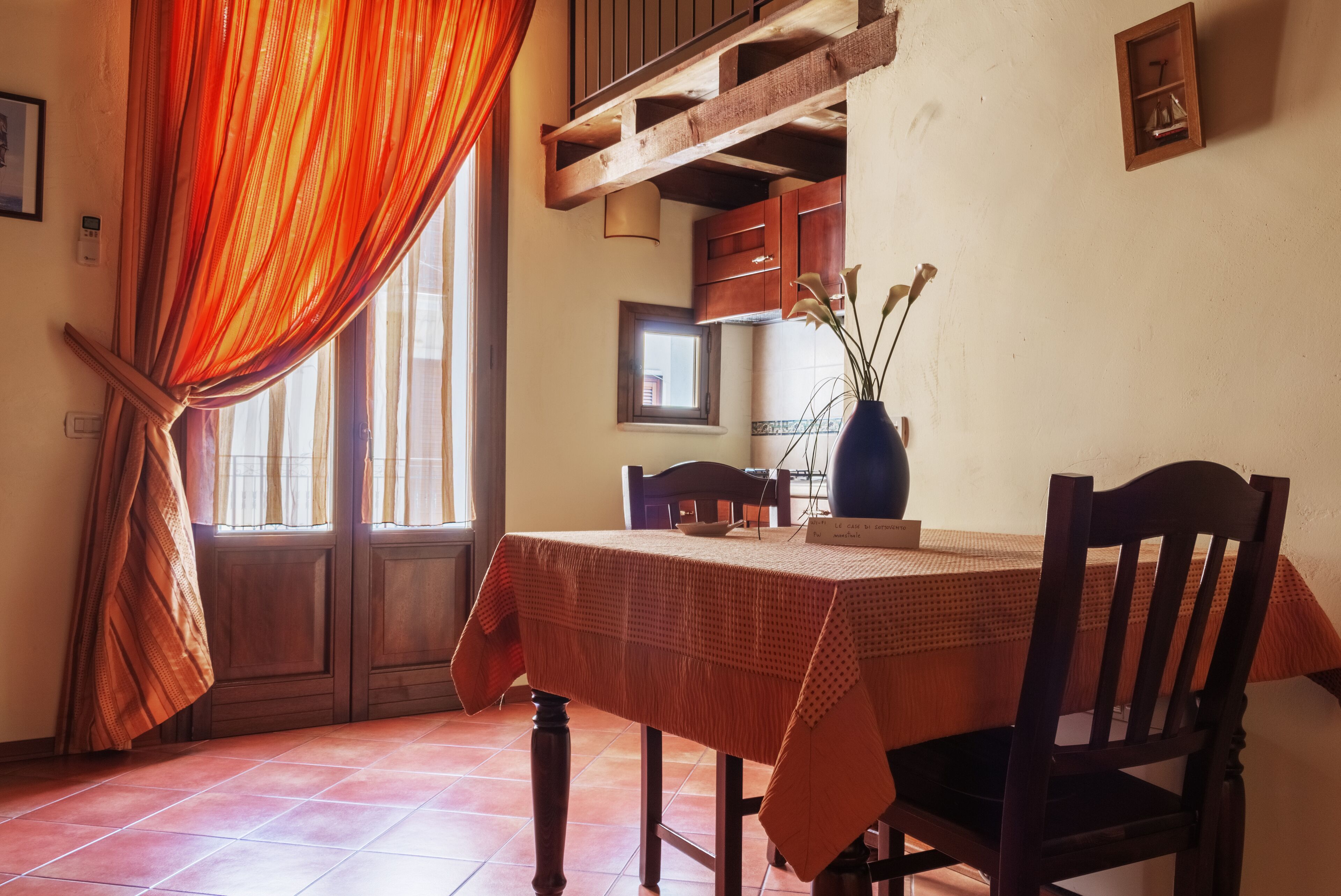 Un comedor con una mesa dispuesta para dos personas, sillas de madera, un florero con flores y una ventana con cortinas.