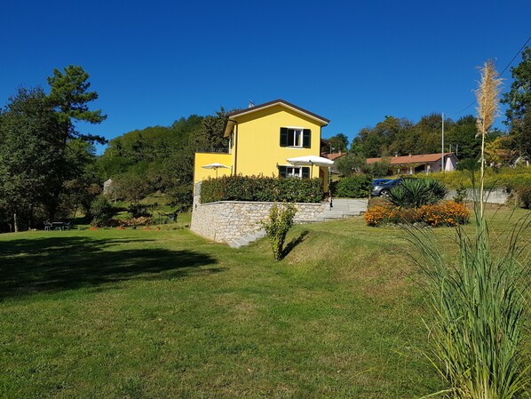 Una casa amarilla con un muro de piedra, rodeada de follaje y un cielo azul despejado.