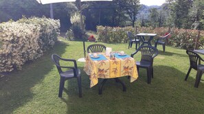 Un área de comedor al aire libre con una mesa dispuesta para dos personas, rodeada de sillas, en un jardín con setos y árboles al fondo.