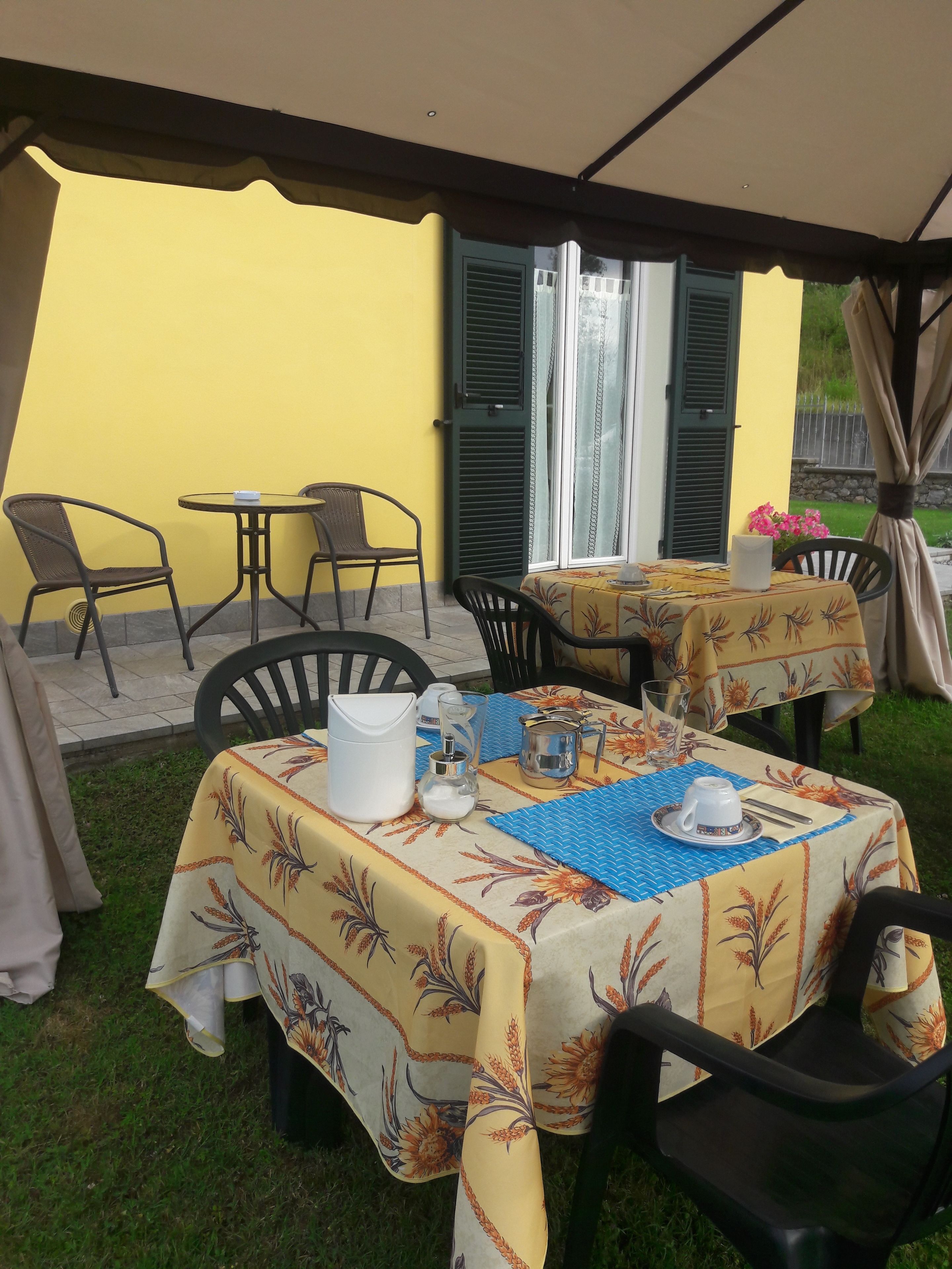 An outdoor patio area with tables and chairs, covered by a canopy, and a yellow wall in the background.