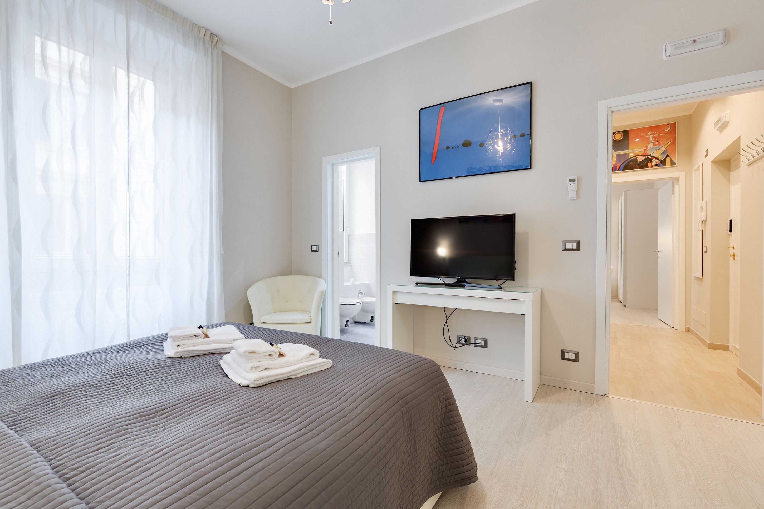 A hotel room with a bed, a TV, a chair, and a bathroom visible through an open door.