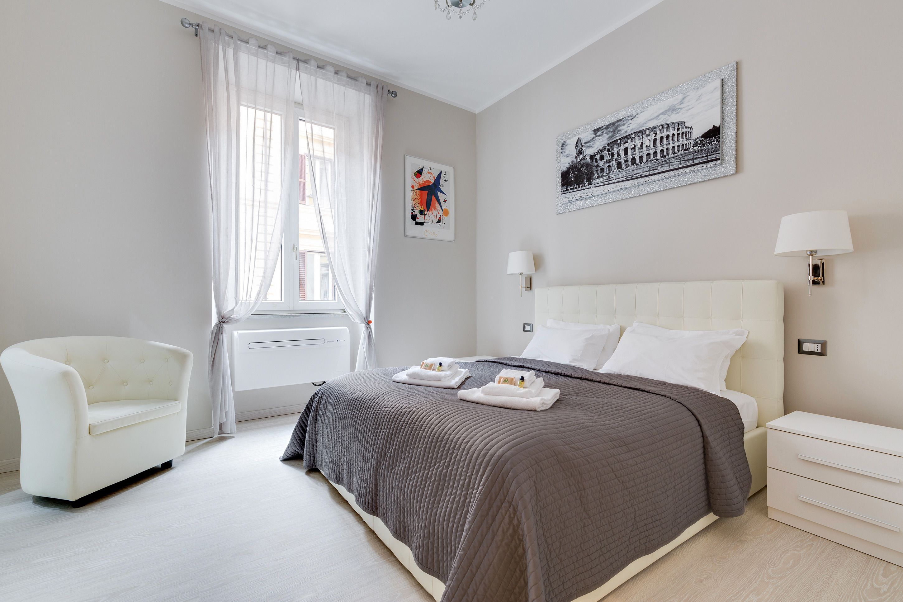 Una habitación de hotel moderna con una cama grande, un sillón blanco y una ventana con cortinas translúcidas.