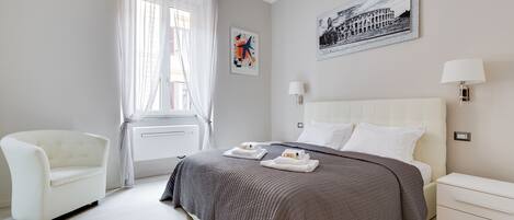 Una habitación de hotel moderna con una cama grande, un sillón blanco y una ventana con cortinas translúcidas.