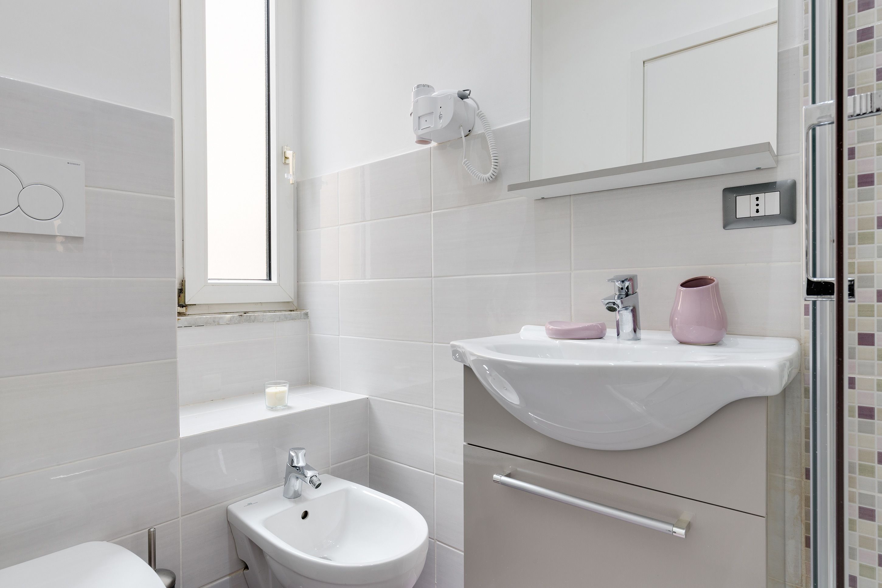 A modern bathroom with a bidet, a sink with a pink vase, and a hairdryer mounted on the wall.