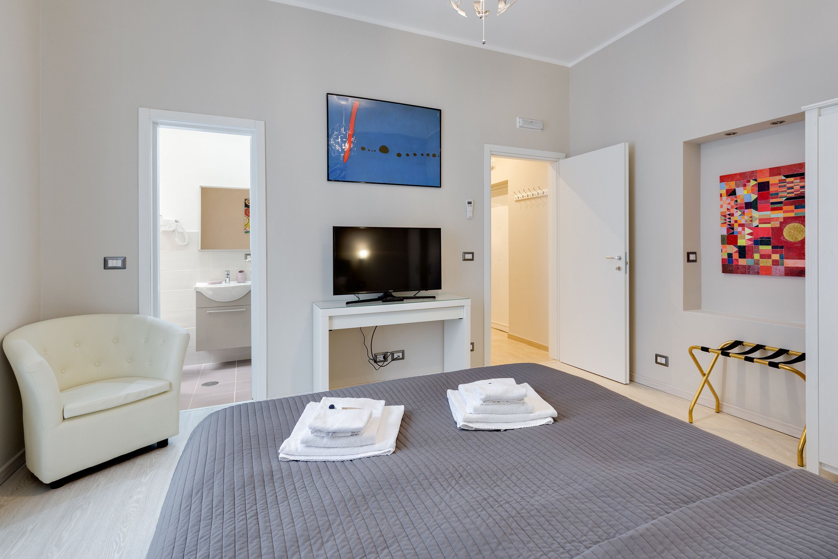 A hotel room with a bed, a TV, a chair, and a bathroom visible through an open door.