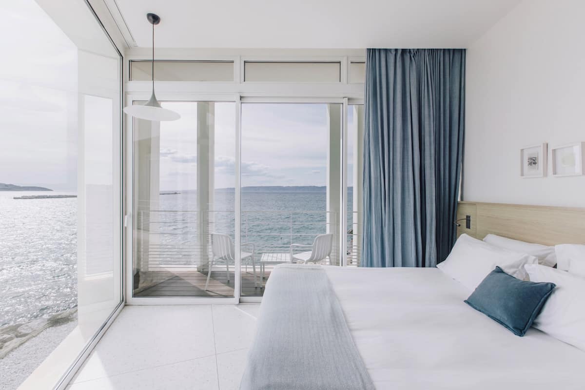 A hotel room with a bed, a view of the sea, and a balcony with chairs.