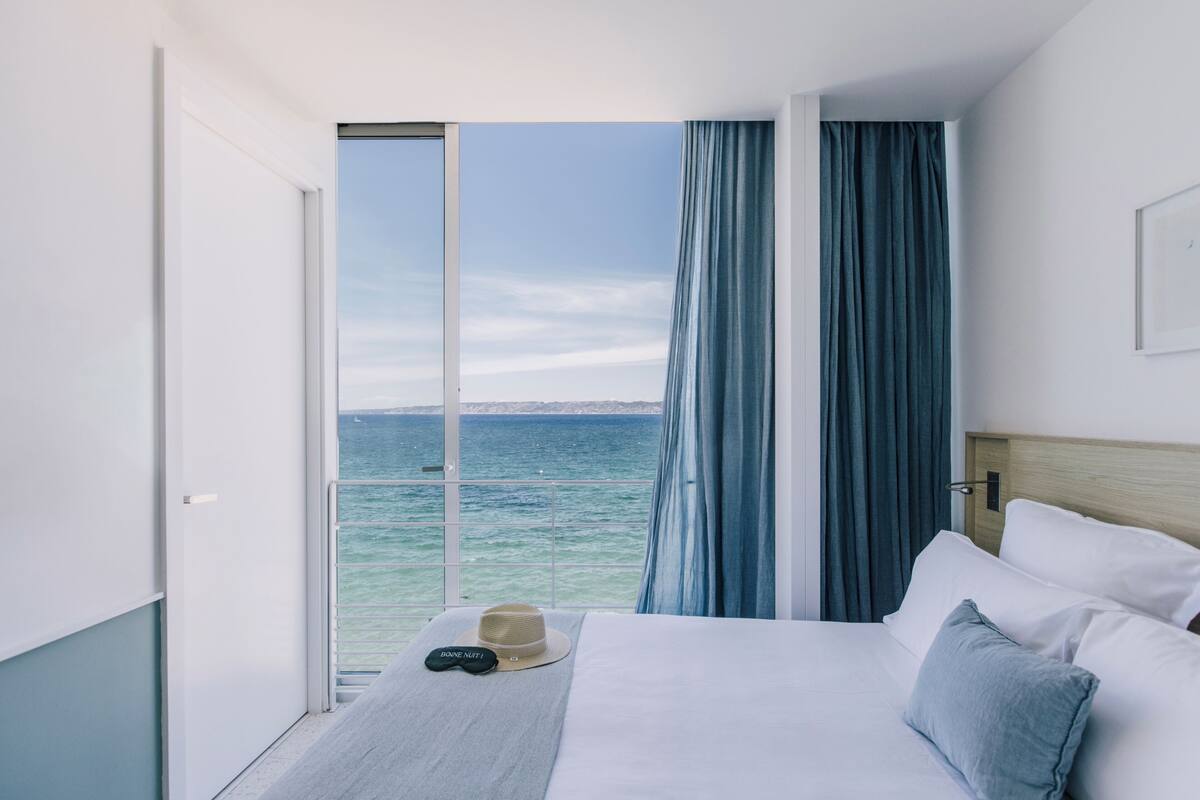 A hotel room with a bed, a hat, a remote control, and a view of the sea through a large window.